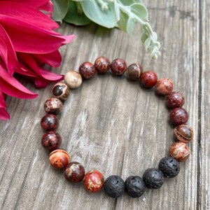 May include: A red, brown and black beaded bracelet with lava rock beads. The bracelet is made with natural stones and is designed to be worn on the wrist.