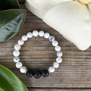 May include: White and black beaded bracelet with lava rock beads. The bracelet is on a wooden surface.