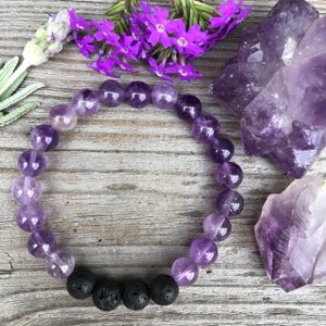 May include: A purple amethyst gemstone bracelet with black lava beads. The bracelet is on a wooden surface with purple flowers and amethyst crystals.