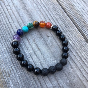 May include: A black beaded bracelet with seven colourful gemstone beads and two lava rock beads. The bracelet is on a wooden surface.