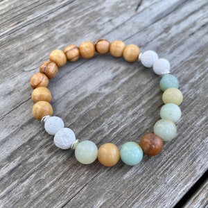 May include: A multi-coloured beaded bracelet with wooden, white and green beads. The bracelet has a gradient colour scheme, starting with brown wood beads, transitioning to white beads, and ending with green beads.