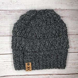 Könnte beinhalten: Eine schwarz-weiß gestrickte Beanie-Mütze mit strukturiertem Muster. Die Mütze hat einen umgeschlagenen Saum und ein kleines, quadratisches, braunes Etikett mit einem Logo auf der Vorderseite. Die Mütze ist aus weichem, warmem Material.