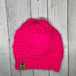 Könnte beinhalten: Eine leuchtend pinkfarbene Häkelmütze mit einem braunen Lederanhänger. Der Anhänger hat ein weißes Totenkopf- und Knochenkreuz-Design.
