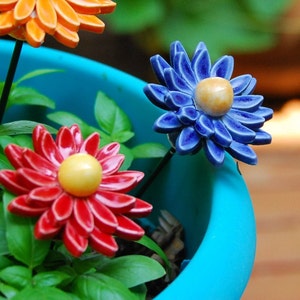 K&ouml;nnte beinhalten: Drei Keramik-Blumen-Dekorationen in einem blauen Topf. Die Blumen sind rot, orange und blau mit gelben Zentren. Die Blumen sind auf Drahtstangen.