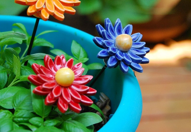K&ouml;nnte beinhalten: Drei Keramik-Blumen-Dekorationen in einem blauen Topf. Die Blumen sind rot, orange und blau mit gelben Zentren. Die Blumen sind auf Drahtstangen.