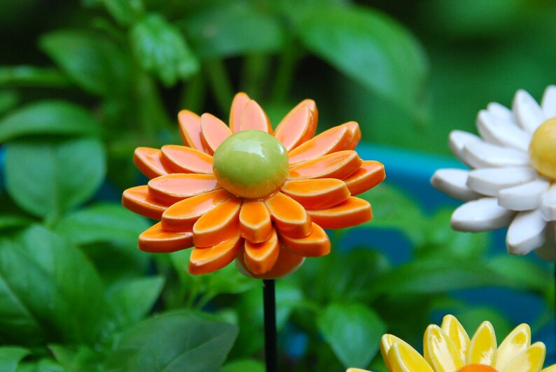K&ouml;nnte beinhalten: Drei Keramik-Blumen-Dekorationen. Die erste Blume ist orange mit einer gr&uuml;nen Mitte. Die zweite Blume ist wei&szlig; mit einer gelben Mitte. Die dritte Blume ist gelb mit einer orangenen Mitte.