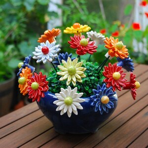 K&ouml;nnte beinhalten: Ein blauer Keramiktopf mit einer bunten Anordnung von Keramikblumen. Die Blumen sind in Rot-, Orange-, Gelb-, Wei&szlig;- und Blaut&ouml;nen gehalten. Der Topf steht auf einer Holzoberfl&auml;che.