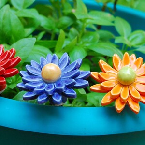 K&ouml;nnte beinhalten: Vier Keramik-Blumentopf-Markierungen in einem blauen Topf. Die Markierungen sind blumenf&ouml;rmig mit roten, blauen, orangefarbenen und gelben Bl&uuml;tenbl&auml;ttern. Die Blumen haben gelbe, gr&uuml;ne und wei&szlig;e Zentren.