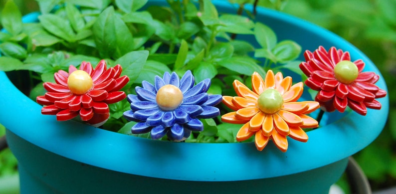 K&ouml;nnte beinhalten: Vier Keramik-Blumentopf-Markierungen in einem blauen Topf. Die Markierungen sind blumenf&ouml;rmig mit roten, blauen, orangefarbenen und gelben Bl&uuml;tenbl&auml;ttern. Die Blumen haben gelbe, gr&uuml;ne und wei&szlig;e Zentren.