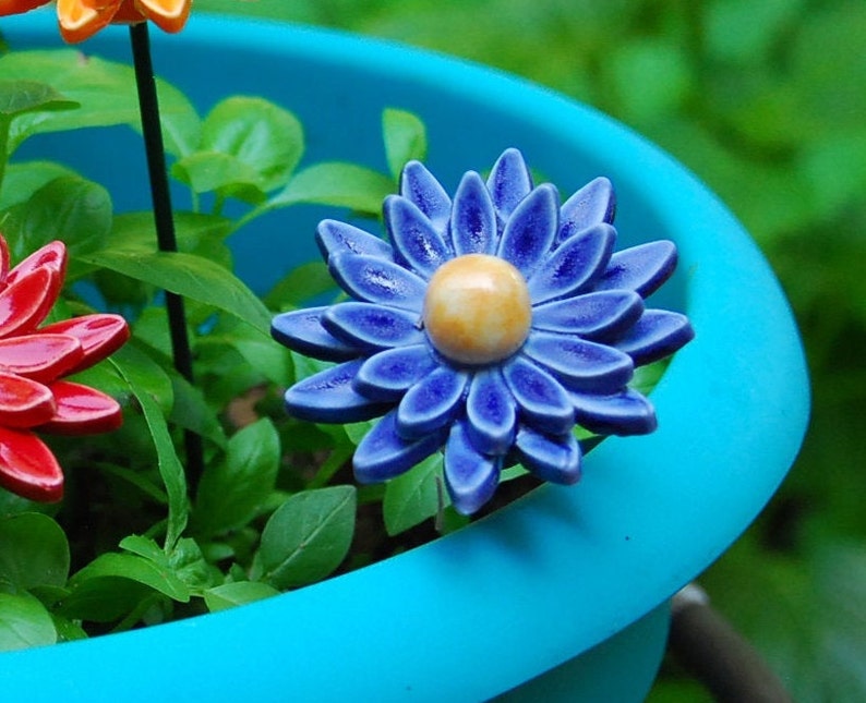 K&ouml;nnte beinhalten: Eine blaue Keramikblume mit gelbem Zentrum sitzt in einem t&uuml;rkisfarbenen Topf mit gr&uuml;nem Laub.