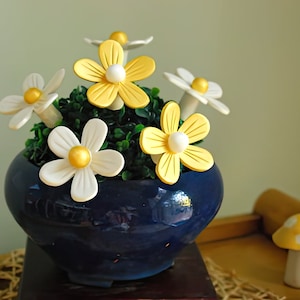 May include: A blue ceramic planter with a white and yellow flower arrangement. The flowers are made of fabric and have a daisy shape. The planter is sitting on a wooden surface.