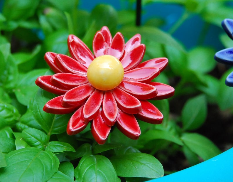 K&ouml;nnte beinhalten: Eine rote Keramikblume mit gelber Mitte, umgeben von gr&uuml;nen Bl&auml;ttern.