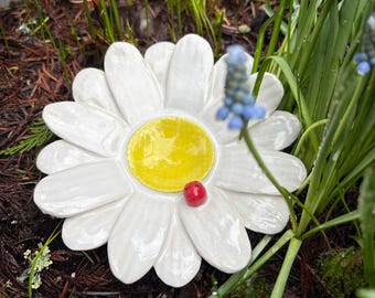 Könnte beinhalten: Eine weiße Keramik-Gänseblume mit gelbem Zentrum und einem kleinen roten Akzent. Die Blume ist von grünen Pflanzen und braunem Mulch umgeben, wodurch eine gartenähnliche Umgebung entsteht. Die Gänseblume ist ein dekoratives Gartenelement.