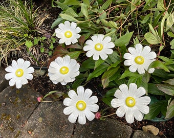 Könnte beinhalten: Eine Ansammlung von sechs weißen, gänseblümchenförmigen Gartenornamenten aus Keramik mit leuchtend gelben Mitten. Jede Blume weist ausgeprägte, abgerundete Blütenblätter in kreisförmiger Anordnung auf, platziert zwischen grünem Laub und dunkler Erde.