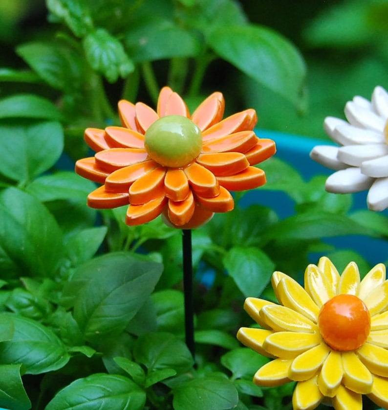K&ouml;nnte beinhalten: Drei Keramik-Blumen-Gartenstecker. Die Blumen sind orange, gelb und wei&szlig;. Die orange Blume hat eine gr&uuml;ne Mitte. Die gelbe Blume hat eine orange Mitte. Die wei&szlig;e Blume hat eine gelbe Mitte.