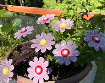 Könnte beinhalten: Eine Ansammlung dekorativer Gartenstecker in Form von hellvioletten Gänseblümchen mit leuchtend roten oder gelben Zentren. Diese Blumenornamente sind in einem runden, terrakottafarbenen Pflanzgefäß mit Erde und grünen Pflanzen angeordnet.