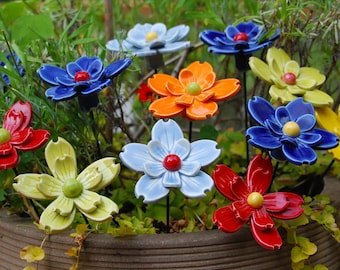 May include: A collection of colorful ceramic flowers with different colored centers. The flowers are arranged in a pot with green foliage.