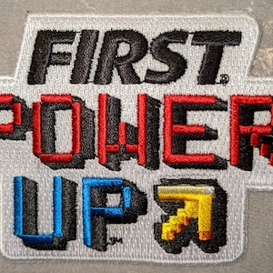 May include: A white embroidered patch with the text "FIRST POWER UP" in black, red, and blue. The "UP" is in a pixelated style with a yellow pixelated image to the right.