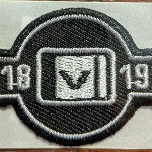 May include: A black and silver embroidered patch with a white V inside a square and the numbers 18 and 19 on either side.