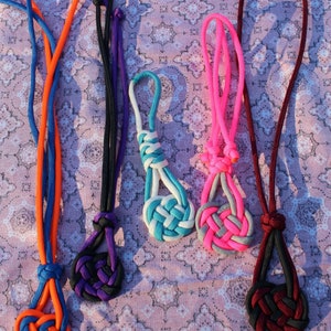 May include: Five paracord Celtic heart knots in different colors, including blue, orange, purple, pink, and red. The knots are tied on different lengths of cord and are displayed on a patterned fabric background.