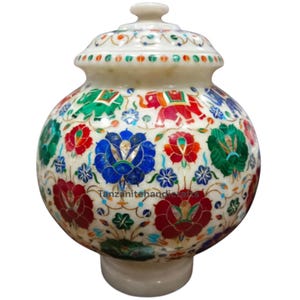 Könnte beinhalten: Dekorative weiße Marmorvase mit Deckel, verziert mit aufwendigen Blumen- und Elefantenmotiven, die mit bunten Edelsteinen eingelegt sind. Die Vase hat einen runden Körper und einen kurzen, zylindrischen Sockel.