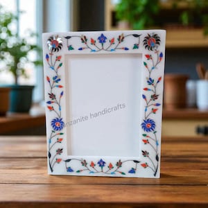 Könnte beinhalten: Weißer Marmor-Bilderrahmen mit floralen Einlegearbeiten. Der Rahmen hat eine rechteckige Öffnung und ist mit einem farbenfrohen Blumenmuster in Rot, Blau und Grün verziert. Der Text "zanzanite handicrafts" ist sichtbar.