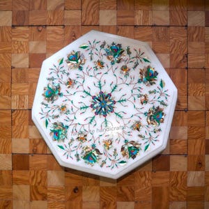Könnte beinhalten: Achteckige Tischplatte aus weißem Marmor mit eingelegtem Blumendesign. Das Design weist türkisfarbene, goldene und orangefarbene Akzente auf. Die Tischplatte steht vor einem Hintergrund aus Holzparkett. Der Text "Tanzanite handicrafts" ist sichtbar.