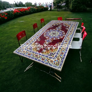 May include: A rectangular table with a red, white and blue patterned inlay. The table is made of marble and has a white border. The table is surrounded by red chairs.