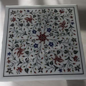 Könnte beinhalten: Eine quadratische Tischplatte aus weißem Marmor mit einem aufwendigen floralen Design, das mit bunten Steinen eingelegt ist. Das Design zeigt rote, blaue, grüne und gelbe Steine. Der Text "Tanzanite handicrafts" ist in der unteren Mitte der Tischplatte sichtbar.