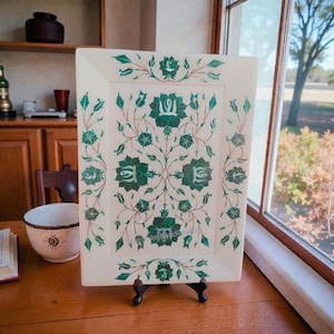 May include: A rectangular off-white stone tray featuring an intricate inlay design of dark green floral motifs and vines. The surface is decorated with symmetrical flower patterns and delicate stems, supported by a small black metal display stand.