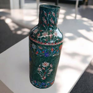 Könnte beinhalten: Eine grüne Marmorvase mit floralen Einlegearbeiten. Die Vase hat eine flaschenähnliche Form mit breitem Boden und schmalem Hals. Das florale Design umfasst weiße, rote und blaue Akzente. Der Text "Tanzanite Handicraft" ist auf der Vase sichtbar.