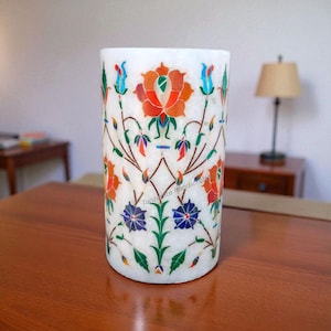 Puede incluir: Jarrón cilíndrico de mármol blanco con incrustaciones florales intrincadas en tonos naranja, azul, verde y rojo. El jarrón presenta un patrón detallado de flores y hojas, mostrando un diseño artesanal. El jarrón mide aproximadamente 15 cm de alto.