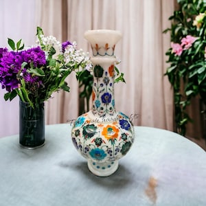 May include: White marble vase with intricate floral designs in blue, green, orange, and yellow. The vase has a round base and a narrow neck. It is sitting on a light blue table.