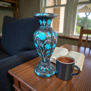 Könnte beinhalten: Eine schwarze Vase mit türkisfarbenen Blumenmustern und silbernen Akzenten. Die Vase steht auf einem Holztisch neben einer schwarzen Tasse mit dunkler Flüssigkeit und einem offenen Buch. Die Vase hat einen breiten Sockel und einen ausgestellten oberen Rand.