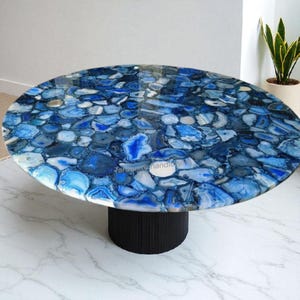 Könnte beinhalten: Ovaler Couchtisch mit einer Mosaikplatte aus blauen und weißen Achatsteinen. Der Tisch hat ein schwarzes, zylindrisches Gestell. Die Oberfläche reflektiert das Licht und steht auf einem weißen Marmorboden.