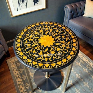 Könnte beinhalten: Ein runder Beistelltisch mit schwarzer Tischplatte und goldenem floralen Intarsienmuster. Der Tisch hat ein goldfarbenes Gestell und einen zentralen Sockel. Der Text "Tanzanite handicrafts" ist auf der Tischoberfläche zu sehen.