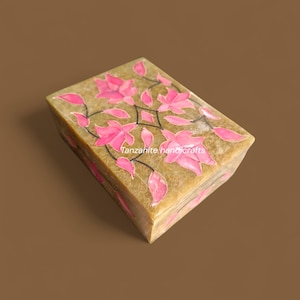 Könnte beinhalten: Eine rechteckige, beige Steinbox mit floralem Design. Rosa Blüten und Blätter sind auf dem Deckel eingelegt, verbunden durch schwarze Linien. Der Text "Tanzanite handicrafts" ist auf der Seite der Box sichtbar.