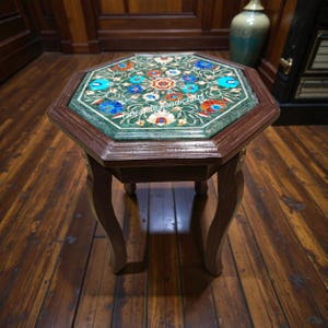 Könnte beinhalten: Ein achteckiger Beistelltisch mit dunklem Holzrahmen und Beinen. Die Tischplatte hat eine grüne Marmoreinlage mit einem floralen Mosaikdesign in Blau-, Rot- und Weißtönen. Der Tisch hat einen Durchmesser von etwa 61 cm.