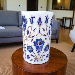 Könnte beinhalten: Eine weiße Marmorvase mit detaillierten blauen floralen Einlegearbeiten. Die Vase hat eine zylindrische Form und ist mit einem sich wiederholenden Muster aus blauen Blumen und Blättern verziert. Der Text "Tanzanite handicraft" ist auf der Vase zu sehen.