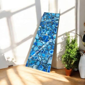 Könnte beinhalten: Eine rechteckige Tischplatte mit einem Mosaikdesign aus blauen und weißen Achatsteinen. Die Oberfläche ist poliert und reflektiert das Licht, mit dem Text "Tanzanite handicrafts" an der Seite. Die Tischplatte lehnt an einer weißen Wand, mit Sonnenlicht und Schatten.