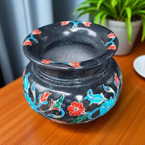 Könnte beinhalten: Eine schwarze Steinvase mit breiter Öffnung und rundem Körper. Sie ist mit roten, blauen und weißen Blumenmustern verziert. Der Text "Tanzanite handicrafts" ist auf der Seite der Vase zu sehen.