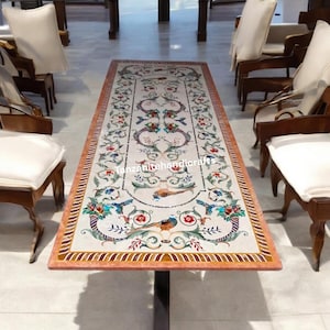 May include: A rectangular dining table with a white marble top featuring an intricate floral design inlaid with colourful stones. The table has a black metal base and is surrounded by chairs.
