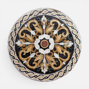 Könnte beinhalten: Ein kreisförmiges Steinmosaik-Medaillon mit einem zentralen braunen Kreis, umgeben von weißen, goldenen und schwarzen floralen Rankenmustern. Der äußere Rand zeigt ein geflochtenes Seildesign in Gold und Schwarz. Der Text Tanzanite handicrafts ist sichtbar.