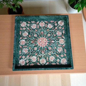 May include: A square, green marble tray with a floral inlay design. The inlay features white, red, and pink flowers arranged in a symmetrical pattern. The tray has a dark border and the text "Tanzanite handicrafts".