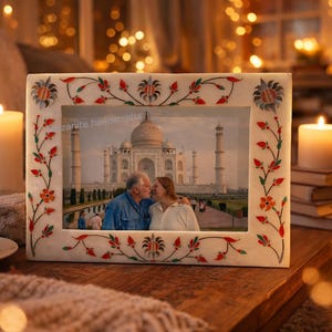 Könnte beinhalten: Ein rechteckiger, cremefarbener Bilderrahmen mit floralen und Blattmustern in Rot, Orange und Grün. Der Rahmen zeigt ein Foto eines Paares, das sich vor dem Taj Mahal küsst. Eine brennende Kerze und eine Kaffeetasse sind zu sehen.