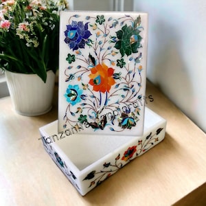 Könnte beinhalten: Weiße Marmorbox mit einem floralen Design, das mit bunten Steinen eingelegt ist. Die Box ist geöffnet und das Innere ist ebenfalls mit einem floralen Design verziert. Der Text "Tanzania Handicrafts" ist auf der Box sichtbar.