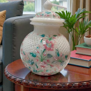 Könnte beinhalten: Eine weiße Marmorvase mit Deckel, verziert mit rosa, grünen und blauen Blumenmustern. Die Vase hat ein gitterartiges Muster und steht auf einem Holztisch. Die Vase ist etwa 25 cm hoch.
