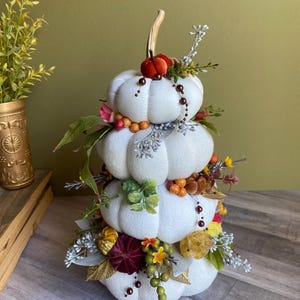 May include: A decorative, stacked pumpkin centerpiece. Three white fabric pumpkins are adorned with fall foliage, berries, and flowers in shades of orange, yellow, and burgundy. A small, red pumpkin tops the arrangement, with a curved, tan stem.