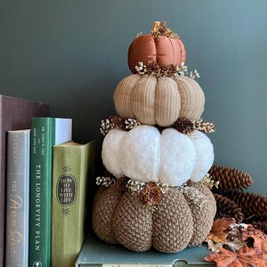 May include: A decorative pumpkin stack in shades of brown, tan, and white, adorned with small floral accents. The top pumpkin is a burnt orange color. The stack is displayed with books and pinecones, creating a fall-themed display.