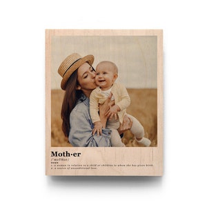 May include: A photo of a mother and child in a field. The mother is wearing a straw hat and a blue denim jacket. The child is wearing a white shirt and is being held by the mother. The photo is printed on a wooden plaque with the word "Mother" and its definition printed below the photo.